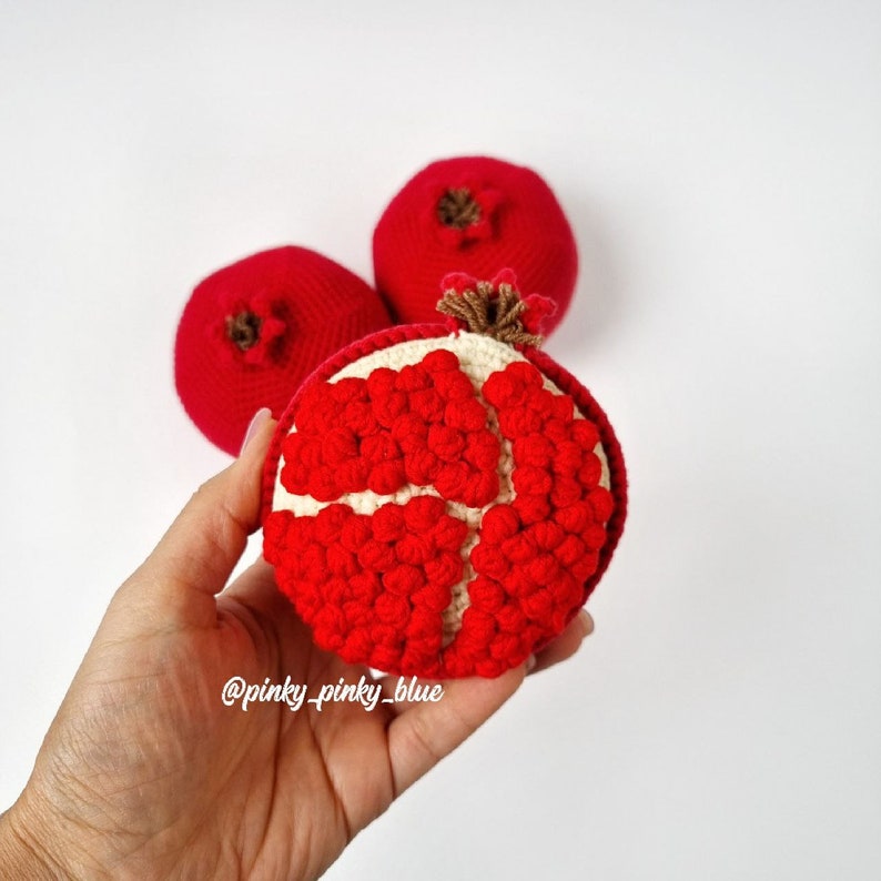May include: Three crocheted pomegranates, one with a white center and red seeds, the other two are solid red. The pomegranate with the white center is being held in a hand. The image includes the text "@pinky_pinky_blue".