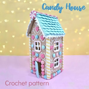 Puede incluir: Casa de caramelo tejida a crochet con un techo azul, detalles de caramelo rosa y blanco y un bastón de caramelo en la parte superior. La casa está sobre una superficie rosa. Patrón de crochet.