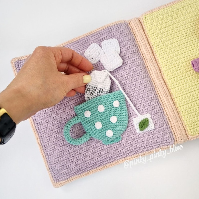 May include: A crocheted teacup with white polka dots, a tea bag, and a small pile of white crocheted tea bags. The teacup is on a purple crocheted background. The text '@pinky_pinky_blue' is in the bottom right corner of the image.