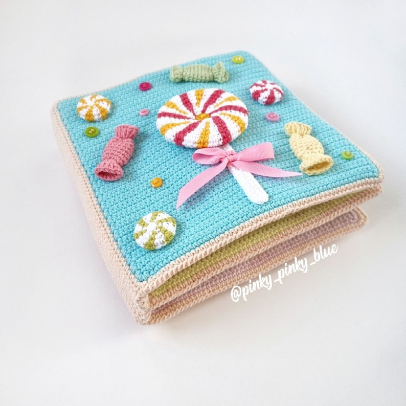 May include: A multi-layered crocheted baby book with a blue cover featuring a variety of colorful crocheted candy shapes, including a lollipop, candy canes, and hard candies. The book is decorated with small white beads and the text "@pinky_pinky_blue".