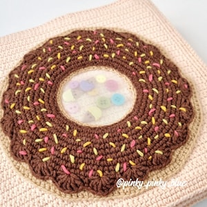 May include: A crocheted donut pillow with a brown center and pink, yellow, and brown sprinkles. The donut has a clear plastic window in the center, revealing colorful buttons.