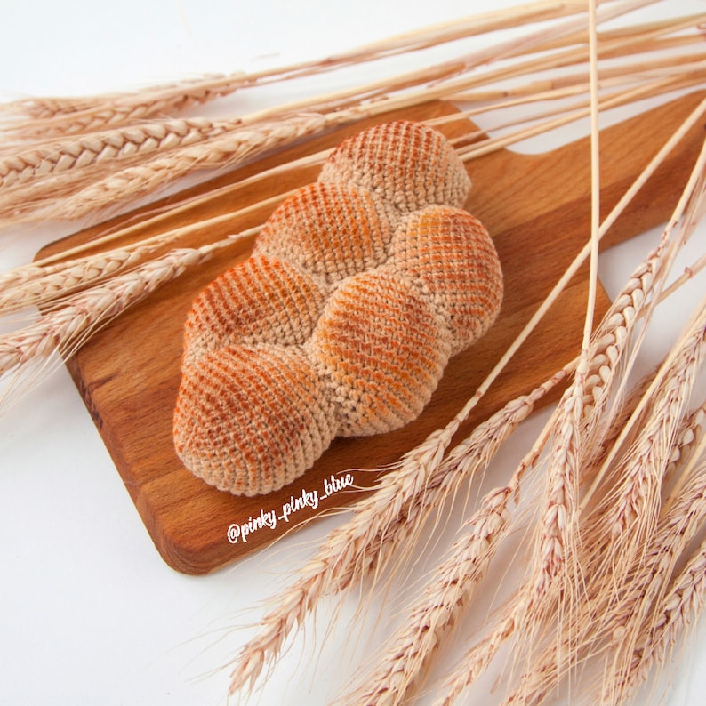 Op de afbeelding: Een gehaakt brood in lichtbruine kleur, dat lijkt op een gevlochten challah, wordt getoond op een houten snijplank omringd door tarwehalmen.
