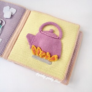 May include: A crocheted baby book with a purple teapot on a yellow page. The teapot has a flame underneath it. The book has a pink and peach border.
