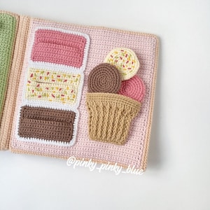 May include: A pink crocheted book with a brown, white, and pink ice cream cone on the front. The ice cream cone has three scoops of ice cream: brown, white with sprinkles, and pink. The book has the text "@pinky_pinky_blue" on the bottom.