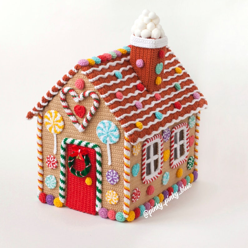 May include: A crocheted gingerbread house with a white frosting roof, candy cane trim, and colorful candy decorations. The house has a red door with a green wreath and two windows.