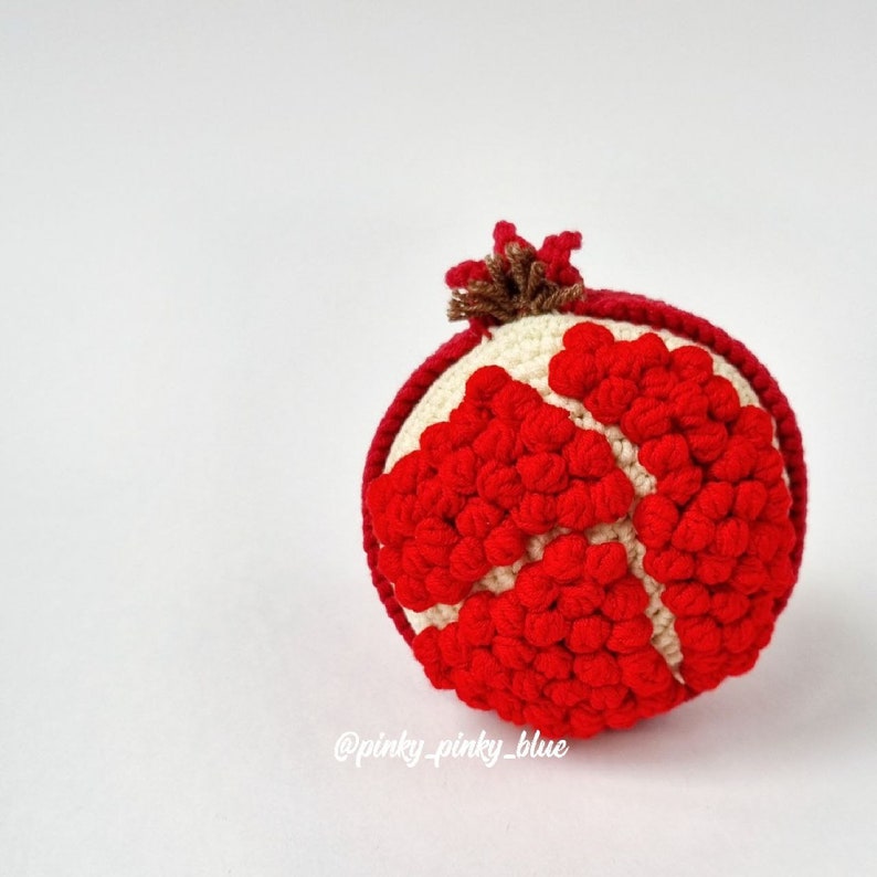 May include: A crocheted pomegranate with red and white yarn. The pomegranate is round and has a brown stem on top.