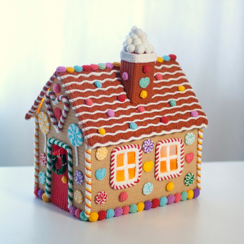 May include: A crocheted gingerbread house with a white frosting roof, candy cane trim, and colorful candy decorations. The house has two windows with lit-up orange lights inside.