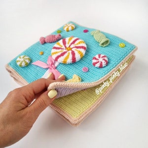 May include: A crocheted book with a candy theme. The cover is blue with a pink and white lollipop, a pink bow, and various candy shapes. The inside of the book is a light green color.