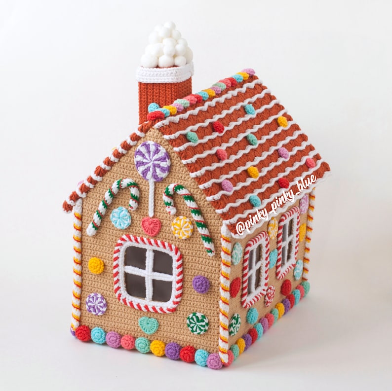 May include: A crocheted gingerbread house with a white frosting roof, candy cane trim, and colorful candy decorations. The house has a window and a chimney with a white frosting top. The house is decorated with the text "@pinky_pinky_blue".