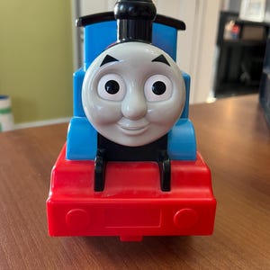 以下が含まれることがあります： 笑顔の顔、青いボディ、赤いベースが特徴の玩具列車。列車は黒いアクセントと黒い煙突が付いています。顔には大きくて丸い目があり、フレンドリーな表情をしています。クラシックな子供のおもちゃ。