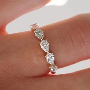May include: A gold ring featuring a series of pear-shaped diamonds. The diamonds are set in a row, creating a sparkling and elegant design. The ring is shown on a finger, highlighting its delicate and refined appearance.