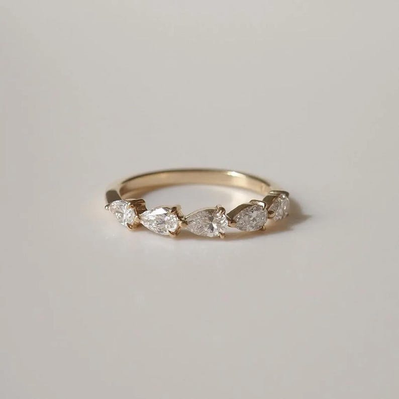 May include: A gold band ring featuring seven pear-shaped diamonds. The diamonds are set in a row, creating a delicate and elegant design. The ring is set against a white background, highlighting the sparkle of the gemstones.