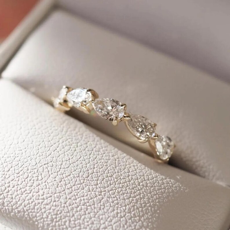 May include: A gold ring with multiple clear, oval-shaped gemstones set in a row. The ring is displayed inside a white, textured jewelry box.