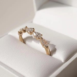 May include: A gold ring with five pear-shaped diamonds set in a row. The ring is displayed inside a white jewelry box, showcasing the ring's design and sparkle. The ring's band is a warm gold color.