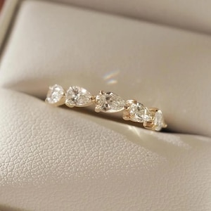 May include: A gold ring featuring a series of pear-shaped, clear gemstones. The ring is set in a gold band and is displayed in a neutral-toned jewelry box. The gemstones are evenly spaced along the band, creating a continuous design.