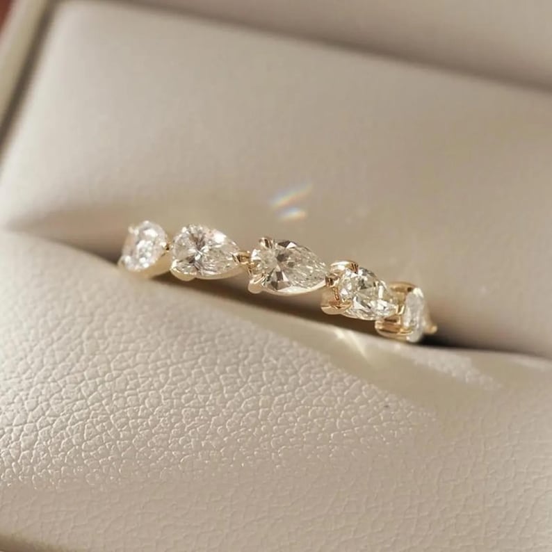 May include: A gold ring featuring a series of pear-shaped, clear gemstones. The ring is set in a gold band and is displayed in a neutral-toned jewelry box. The gemstones are evenly spaced along the band, creating a continuous design.