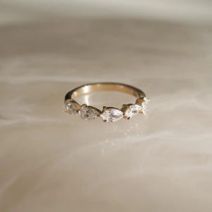 May include: A gold ring featuring a series of pear-shaped, clear gemstones. The ring's band is a warm gold color, and the gemstones are set closely together, creating a continuous sparkle. The ring is displayed on a light-colored surface.