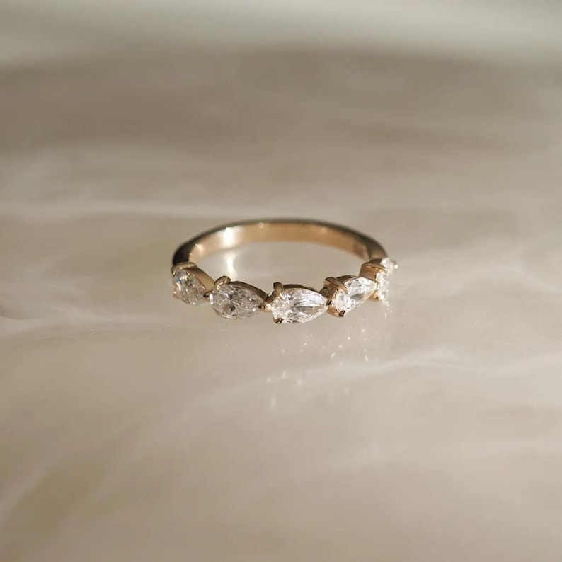 May include: A gold ring featuring a series of pear-shaped, clear gemstones. The ring's band is a warm gold color, and the gemstones are set closely together, creating a continuous sparkle. The ring is displayed on a light-colored surface.