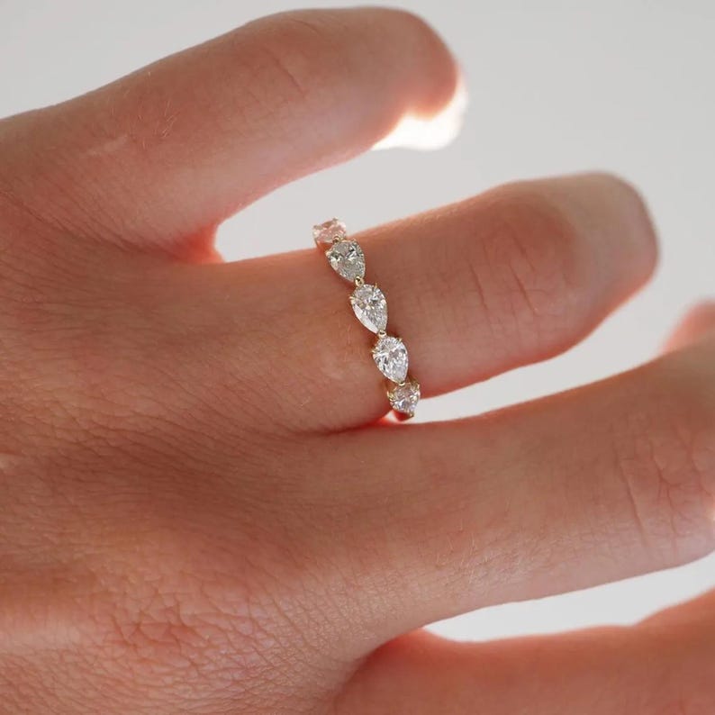 May include: A gold ring featuring a series of pear-shaped diamonds. The diamonds are set in a row, creating a sparkling and elegant design. The ring is shown on a finger, highlighting its delicate and refined appearance.