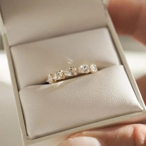May include: A gold ring with a series of clear, pear-shaped gemstones set in a row. The ring is displayed inside a white jewelry box with a soft, textured interior. The ring's design is elegant and minimalist.