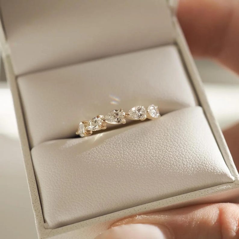 May include: A gold ring with a series of clear, pear-shaped gemstones set in a row. The ring is displayed inside a white jewelry box with a soft, textured interior. The ring's design is elegant and minimalist.