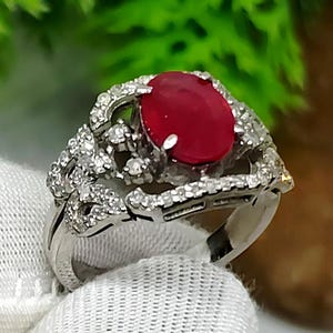 Puede incluir: Un anillo de plata con un gran rubí de corte ovalado. El rubí es de color rojo intenso y está rodeado de piedras preciosas más pequeñas y transparentes. El anillo tiene un diseño ornamentado y detallado con un acabado pulido.