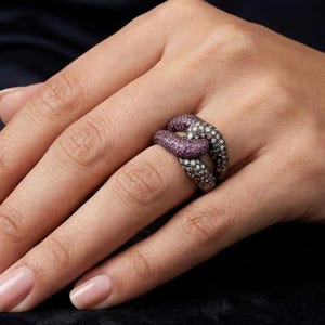 Può includere: Primo piano di un anello dal design unico. L'anello presenta due fasce intrecciate, una ornata di piccole pietre bianche scintillanti e l'altra di vibranti pietre viola. L'anello è posto su uno sfondo scuro.