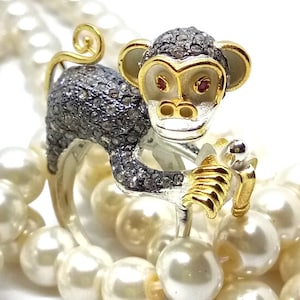 Könnte beinhalten: Ein Ring aus Silber und Gold in Form eines Affen mit schwarzen Diamanten und roten Rubinaugen. Der Affe hält eine goldene Banane in seiner Hand. Der Ring liegt auf einem Bett aus weißen Perlen.