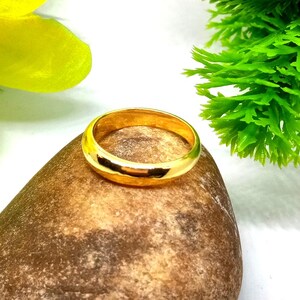 以下が含まれることがあります： 滑らかで磨かれた表面を持つ金色の指輪。茶色の石の上に置かれ、背景には緑の葉と黄色の花びらがあります。指輪のデザインはシンプルでエレガントです。