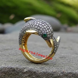 May include: A gold and silver dolphin ring with a green gem eye. The dolphin's body is covered in small, sparkling stones. The ring is set on a gray surface with a blurred green background.