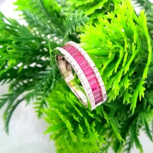 May include: A silver ring with a band of rectangular, pink gemstones and a row of small, clear gemstones. The ring is set against a backdrop of bright green foliage.