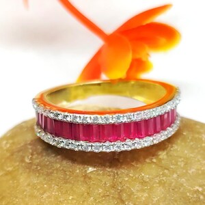 Puede incluir: Un anillo de oro con una banda de piedras preciosas rojas y un borde de pequeñas piedras transparentes. El anillo está sobre una superficie marrón claro, con una flor naranja en el fondo.
