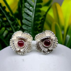 May include: A pair of flower-shaped stud earrings. Each earring features a silver-colored, layered petal design surrounding a round, deep red gemstone center. The earrings are set against a blurred background of green and yellow foliage.