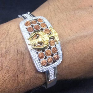 Puede incluir: Pulsera plateada con un elemento central rectangular. Este elemento presenta cuentas redondas marrones, un emblema dorado y está enmarcado por piedras claras y brillantes. La pulsera es una mezcla de tonos plateados y dorados.