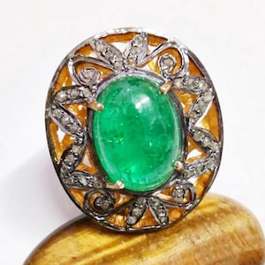Puede incluir: Anillo con una esmeralda verde ovalada. El anillo presenta un engaste decorativo con pequeñas piedras de acento claras y un diseño de filigrana dorada. El anillo está sobre una superficie dorada.