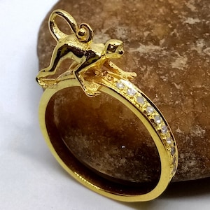 Könnte beinhalten: Ein goldener Ring mit einem Affenanhänger und einer Reihe kleiner, klarer Steine um das Band.