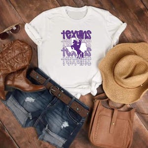 May include: White t-shirt with a purple graphic that says "Texas" with a silhouette of a cowboy on a horse. The image includes a pair of brown cowboy boots, denim shorts, a brown leather purse, and a straw hat.