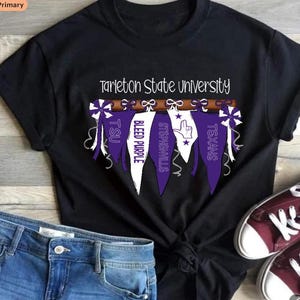 May include: Black t-shirt featuring the text "Tarleton State University." Below the text are purple and white pennants with the words "TSU," "Bleed Purple," "Stephenville," and "Texans." The shirt is paired with blue denim shorts and maroon sneakers.