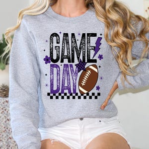 May include: A light gray sweatshirt with the words "GAME DAY" in black and purple, along with a brown football graphic. The design includes a purple lightning bolt and floral accents. The bottom of the design has a black and white checkered pattern.