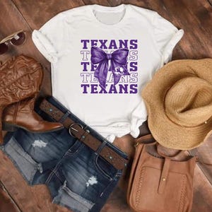 May include: White t-shirt with the word "TEXANS" in purple, featuring a purple bow graphic. The image includes denim shorts, brown leather boots, a straw hat, sunglasses, and a brown leather purse.
