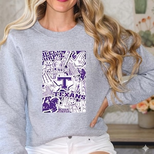 May include: A heather gray crewneck sweatshirt featuring a rectangular purple and white graphic print. The design includes the text Tarleton Texans, Bleed Purple, and various illustrations including a mascot, a hand gesture, and a large letter T.