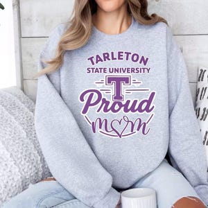 May include: A light gray crewneck sweatshirt featuring purple screen-printed text that reads Tarleton State University Proud mom with a large letter T and a heart integrated into the word mom, centered on the front of the garment.