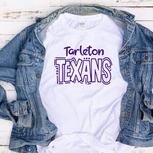 May include: A white t-shirt featuring the purple text Tarleton TEXANS in a stylized font, layered underneath a blue denim jacket. The shirt has a crew neckline and short sleeves, with the graphic centered on the chest area of the garment.
