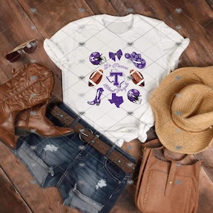May include: A white t-shirt featuring a purple and brown graphic print of football helmets, a bow, a cowboy hat, footballs, a cowboy boot, a map of Texas, and the letter T. The shirt is displayed with brown leather boots, denim shorts, and a straw hat.