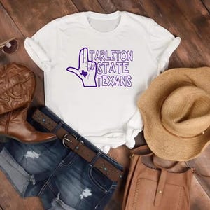 May include: A white short-sleeve t-shirt featuring a purple graphic of a hand making a sign with a small Texas state outline inside, and the text Tarleton State Texans. The shirt is displayed with brown cowboy boots, denim shorts, and a hat.