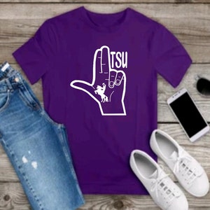 May include: Purple t-shirt with a white hand making the "L" sign with a white horse and rider inside the hand. The text "TSU" is above the hand.