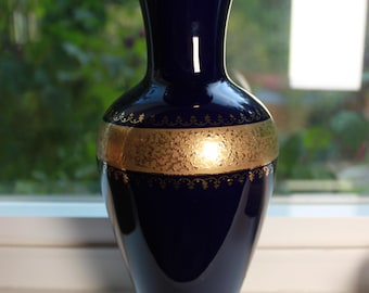 May include: A deep cobalt blue ceramic vase featuring a wide gold band around the centre with an intricate filigree pattern. The vase has a flared rim and a circular base, both finished with a polished gold trim that reflects light.