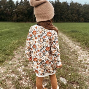 Könnte beinhalten: Ein Kind trägt eine beige Mütze, ein Kleid mit Blumenmuster, braune Strumpfhosen und braune Stiefel mit weißem Fellbesatz. Das Kleid hat eine weiße Basis mit orangefarbenen und cremefarbenen Blumenmustern.