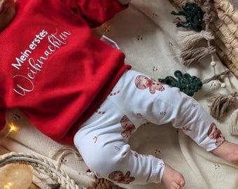 Puede incluir: Una camiseta roja de manga larga con textura acanalada y el texto blanco Mein erstes Weihnachten, junto a un pantalón blanco con un estampado repetitivo de galletas de jengibre y bastones de caramelo. Conjunto infantil de tela suave.