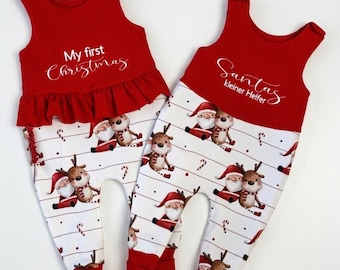 Puede incluir: Dos peleles sin mangas con cuerpos rojos acanalados y pantalones blancos con un estampado de Papá Noel y renos. El pelele de la izquierda tiene el texto My first Christmas y el de la derecha el texto Santas kleiner Helfer en el pecho.
